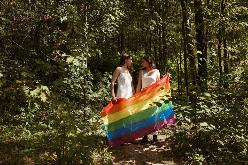 Couple in white dresses holding a rainbow flag in a lush forest setting, symbolizing love and unity.